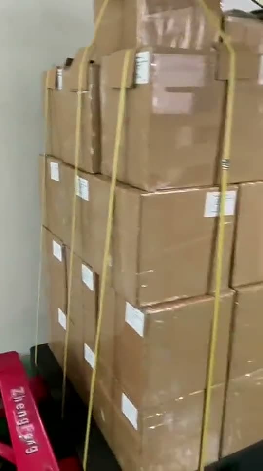 A pallet of kraft cartons in a factory dispatch bay, shrink-wrapped, strapped in yellow banding and carrying printed carton labels, standing on a pallet truck ready to leave.